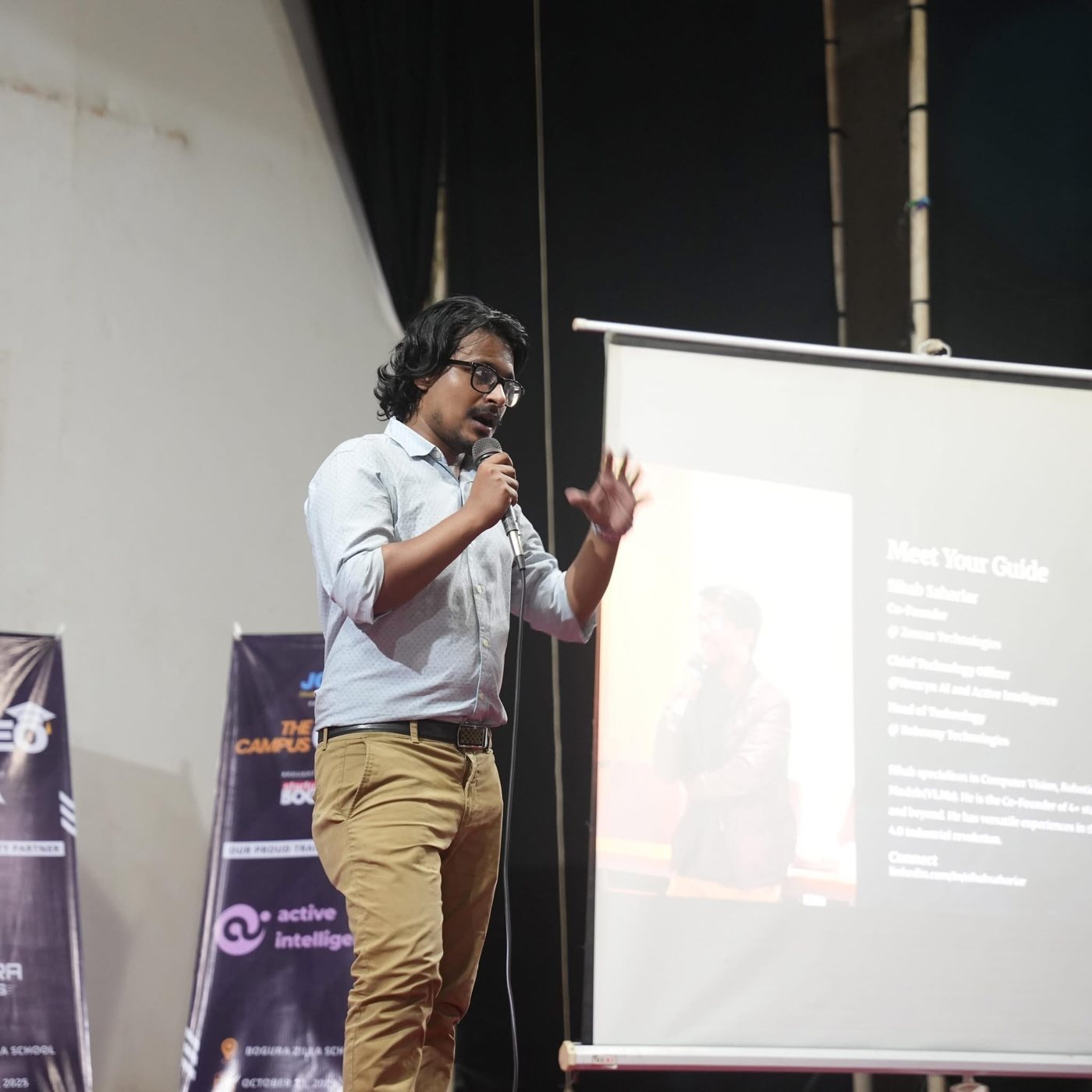 Sihab speaking into a microphone on stage beside a projector screen reading Meet Your Guide